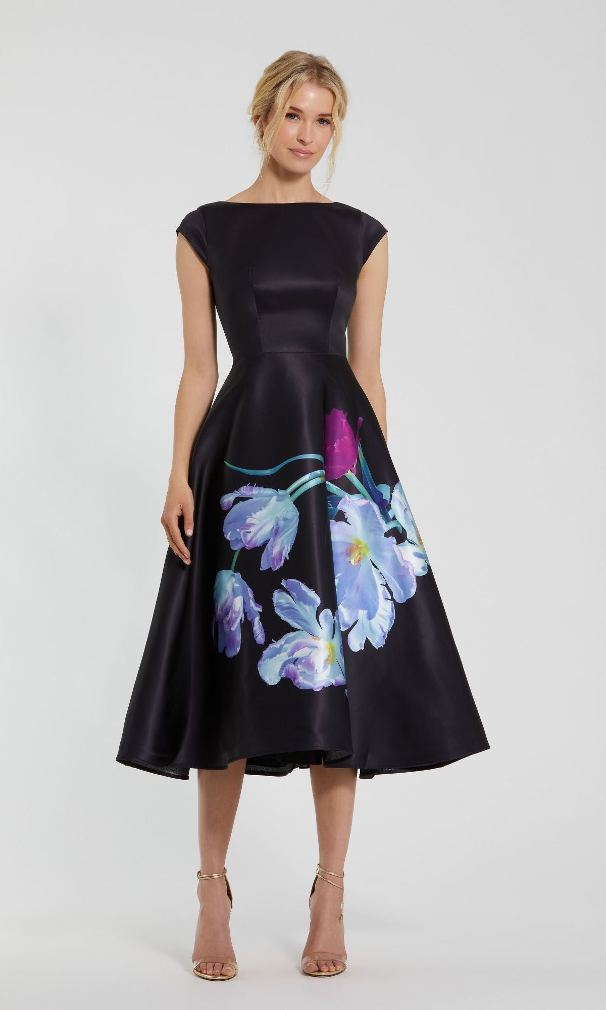 A woman stands against a plain background wearing the Mac Duggal 20995 Black Satin Wedding-Guest Dress, featuring a floral print, cap sleeves, and a mid-length hem.